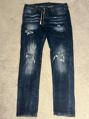 Dsquared2 blå slitna jeans - Säljer ett par mörkblå jeans från Dsquared2 med slitningar, hål och färgstänk för en riktigt edgy look. Jeansen har klassisk femficksmodell, dragkedja och knappgylf samt Dsquared2-lapp bak i midjan. Materialet är robust denim i bomull.