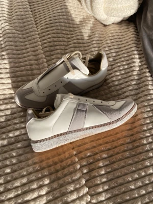 Maison Margiela Gats sneakers beige/vit - Maison Margiela Gats sneakers i beige och vitt med detaljer i mocka och skinn. Klassisk siluett med rund tå, snörning och gummisula. Snygg minimalistisk design och diskret branding på sulan. Perfekta för dig som gillar stilrena och lyxiga sneakers.
