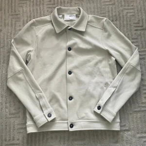 Beige overshirt från Selected Homme - Stilren beige overshirt från Selected Homme med klassisk krage och svarta knappar. Jackan har raka linjer, diskreta sömmar och är tillverkad i ett mjukt bomullsmaterial. Perfekt för lager-på-lager och enkel att matcha med olika outfits. Priset är inte hugget i sten! Vid fler frågor eller funderingar tveka inte på att skriva!