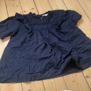 Mörkblå blus med volang från Gina Tricot - Säljer en mörkblå blus från Gina Tricot i storlek 170. Blusen har korta ärmar, volangdetaljer vid axlarna och ett dekorativt band med broderad spets framtill. Materialet är mjukt och lätt bomull, perfekt för en chill och snygg look.