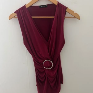 Vinröd omlottopp med pärldetalj från Vila - Säljer en vinröd omlottopp från Vila med djup v-ringning och ärmlös design. Toppen har en snygg drapering framtill och en rund pärlprydd detalj i midjan som ger extra stilpoäng. Perfekt för dig som vill sticka ut med en elegant och trendig look.