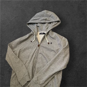 Grå zip hoodie med vita snören från Morris  - Grå hoodie från Morris Company med dragkedja, vita snören med bruna detaljer och broderad logga på bröstet. Tillverkad i mjuk bomull och har huva samt långa ärmar. Perfekt för chill dagar eller när du vill ha en clean och enkel stil. Kan tänka mig gå ner en bit i pris för en snabb affär 😁