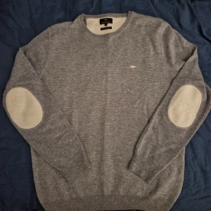 Grå kashmirtröja från Fynch-Hatton - Snygg grå stickad tröja från Fynch-Hatton i mjuk merinoull och kashmir. Tröjan har rund hals, långa ärmar och ljusa armbågslappar som ger en stilren touch. Liten broderad logga på bröstet. Perfekt för kyliga dagar.