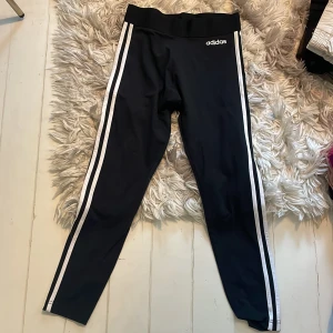Svarta Adidas leggings med vita ränder - Svarta leggings från Adidas med klassiska vita ränder längs sidorna och Adidas-logga på höften. Vanligt tyg material och bred resår i midjan för skön passform. Perfekta för träning eller chill.