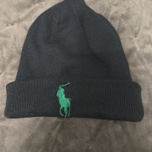 Mörk blå mössa från Polo Ralph Lauren - Mörkblå  ribbstickad mössa från Polo Ralph Lauren med grön broderad logga framtill. Klassisk design med uppvikt kant och mjukt material som håller dig varm. Perfekt accessoar för kyliga dagar och snygg till streetwear.