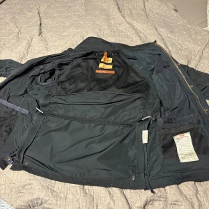 Svart Parajumpers parkajacka med patch - Svart parkajacka från Parajumpers med flera fickor, patch på ärmen och gul detalj vid kragen. Jackan har dragkedja, knappar och meshfoder på insidan. Perfekt för dig som vill ha en funktionell och snygg jacka med streetkänsla.