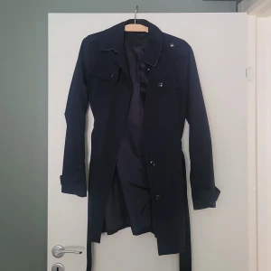 Mörkblå trenchcoat från Tommy Hilfiger - Snygg mörkblå trenchcoat från Tommy Hilfiger i klassisk modell med knappar framtill, bälte i midjan och långa ärmar. Jackan har en stilren design med axelklaffar och detaljer vid ärmsluten. Perfekt för dig som vill ha en tidlös och clean look.