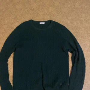 Mörkgrön stickad tröja J.Lindeberg - Snygg mörkgrön stickad tröja från J.Lindeberg i storlek M. Tröjan har ett diskret kabelstickat mönster och rund halsringning. Perfekt för lager-på-lager och passar till både jeans och chinos.