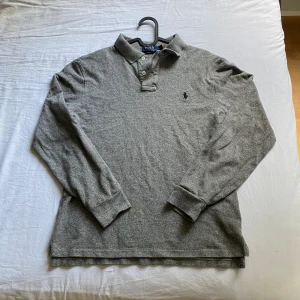Polo Ralph Lauren tröja - Slim fit grå långärmad pikétröja från Polo Ralph Lauren med klassisk krage och knappar framtill. Tröjan har broderad logga på bröstet och är gjord i mjuk bomull. Perfekt för dig som gillar stilren och enkel design. Passar även S