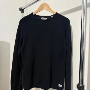 Svart stickad tröja från Jack & Jones - En stilren svart stickad tröja från Jack & Jones i storlek S. Tröjan har rund halsringning och långa ärmar, perfekt för lager-på-lager under kyliga dagar. Enkel design med diskret logga nertill. Passar dig som gillar klassisk och clean stil.