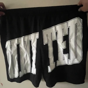 Svarta shorts med stor text från FB Sister - Svarta shorts från FB Sister med elastisk midja och stor vit text 'UNITED' tryckt över hela baksidan. Sköna och loose fit, perfekta för chill dagar eller träning. Materialet känns mjukt och lätt, troligen polyester eller bomullsmix.