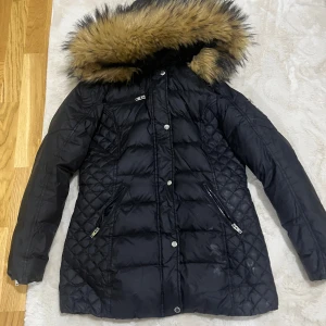 Svart dunjacka Rockandblue med päls - Svart dunjacka från Rockandblue med quiltade detaljer, dragkedjor och knappar framtill. Jackan har en stor luva med fluffig päls och flera fickor med dragkedja. Perfekt för kalla dagar och riktigt snygg passform. Helt äkta