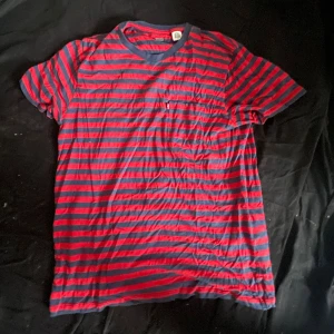 Randig t-shirt från Levi's i rött och blått - Säljer en randig t-shirt från Levi's i rött och mörkblått. T-shirten är i mjuk Supima-bomull, har rund halsringning med blå kant och en liten bröstficka. Perfekt för en chill och avslappnad stil.