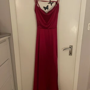 Röd långklänning med snörning i satin - Säljer en röd långklänning i satin med smala axelband och snygg snörning i ryggen. Klänningen har en draperad urringning och öppen rygg med flera band som ger en unik look. Perfekt för en bal. 