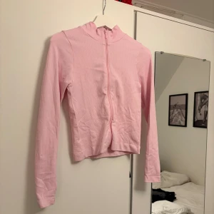 Ljusrosa ribbad tränings ziptröja - Supersöt ljusrosa ribbad tröja med dragkedja hela vägen framtill. Tröjan har lång ärm och en smal passform som sitter snyggt. Perfekt att slänga på sig över ett linne eller t-shirt. Enkel och stilren design som passar till det mesta.