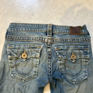 Blå jeans med broderade bakfickor - Snygga blå jeans från True Religion med ikoniska broderade bakfickor och dekorativa knappar. Säljer då de tyvärr inte passar längre. Köpte för ungefär 1000 kr, säljer billigare eftersom dem slitnat lite här och där. 🥰