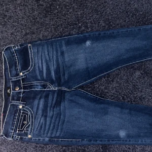 True Religion jeans i mörkblå denim - Säljer ett par snygga True Religion jeans värt 1200 i mörkblå tvätt med kontrasterande vita sömmar och ikoniska bakfickor med lock och silverfärgade knappar. Klassisk femficksmodell och patch bak i midjan. Perfekta för dig som gillar streetwear och detaljer.