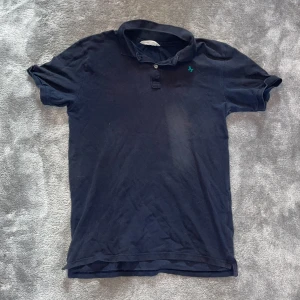 Mörkblå pikétröja från Abercrombie - Klassisk mörkblå pikétröja från Abercrombie & Fitch med liten blå logga på bröstet. Tröjan har krage, korta ärmar och knäppning med två knappar. Tillverkad i 100% bomull för en skön och avslappnad känsla.