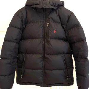Svart dunjacka från Polo Ralph Lauren - Svart dun pufferjacka från Polo Ralph Lauren från Kidsbrandstore. Jackan har huva, dragkedja framtill och tre fickor med dragkedja. Kommer inte med kvitto.