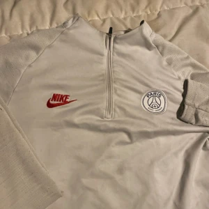 Vit Nike PSG träningströja half zip - Säljer en vit långärmad träningströja från Nike med PSG-logga på bröstet och röd Nike-swoosh. Tröjan har half zip framtill och är tillverkad i ett lätt, syntetiskt material som andas. Perfekt för fotbollsträning eller chill vardagsstil.