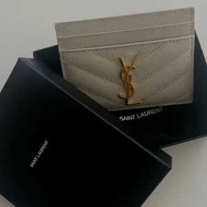 Beige Saint Laurent plånbok i skinn - Supersnygg beige plånbok från Saint Laurent i lyxigt skinn med quiltad design och ikoniska YSL-loggan i guld framtill. Perfekt storlek för kort och enkel att ha med sig i väskan. Kommer med originalask.