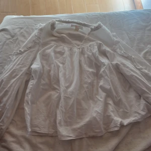 Vit blus med volang från Gina Tricot - Säljer en söt vit blus från Gina Tricot i storlek 146/152. Blusen har långa ärmar och fina volangdetaljer längs axlar och halsringning. Den är tillverkad i mjuk bomull och har en luftig, lite lös passform som ger en chill vibe.