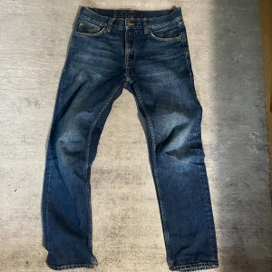 Blå jeans från Nudie Jeans, gritty jackson - Säljer ett par klassiska blå Grim Tim jeans från Nudie Jeans. Modellen har raka ben, fem fickor och snygga slitningar på bakfickorna. Jeansen är tillverkade i 100% bomull och har orangea sömmar samt mässingsfärgade nitar. Perfekta för en avslappnad stil. 39,5cm midja. 72cm innerbenslängd