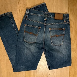 Nudie Jeans  - Snygga blå jeans från Nudie Jeans Co i klassisk tvätt med orange kontrastsömmar och ikonisk brodyr på bakfickorna. Väldigt välbehållna och har inga som helst defekter. Perfekta för dig som gillar stilrena och tidlösa jeans.
