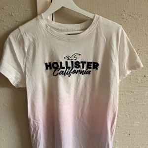 Vit Hollister t-shirt med tryck - Snygg vit t-shirt från Hollister med svart broderat logotyp-tryck och texten 'California' på bröstet. T-shirten har rund hals och korta ärmar, och är gjord i mjuk bomull. Perfekt för en chill och avslappnad stil.