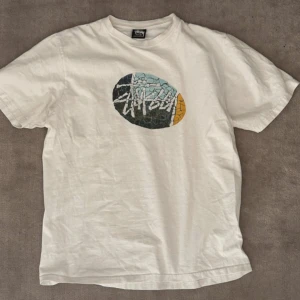 Vit Stüssy t-shirt med grafiskt tryck - Vit t-shirt från Stüssy i bomull med ett stort grafiskt tryck på bröstet i svart, blått och gult. Klassisk passform med rund hals och korta ärmar. Perfekt för dig som gillar streetwear och snygga prints.