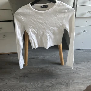 Vit croppad långärmad topp - Säljer en vit croppad långärmad topp. Toppen är ribbad och har en enkel, stilren design med rund halsringning. Perfekt att matcha med jeans eller kjol för en clean look.
