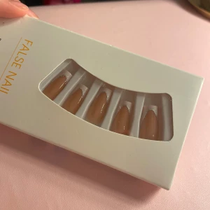 Nude lösnaglar med silverdetalj - Säljer ett set med nude-färgade lösnaglar i mandelform med en tunn silverglittrig linje vid nagelbandet. Paketet innehåller flera storlekar, självhäftande stickers och en rosa nagelfil. Perfekt för dig som vill ha snygga naglar snabbt.