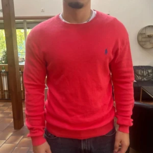 Ralph Lauren stickad tröja - Riktigt snygg röd tröja från Polo Ralph Lauren i slim fit, storlek M. 70cm lång. Tillverkad i mjuk pima bomull och har rund halsringning samt det klassiska blå broderade logotypen på bröstet. Perfekt för dig som gillar stilrena plagg!