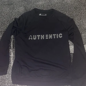 Svart sweatshirt AUTHENTIC Dressmann - Svart sweatshirt från Dressmann med trycket 'AUTHENTIC' i vitt framtill. Tröjan har rund halsringning och långa ärmar, perfekt för en chill och stilren look. Tillverkad i mjuk bomull som känns skön mot huden.