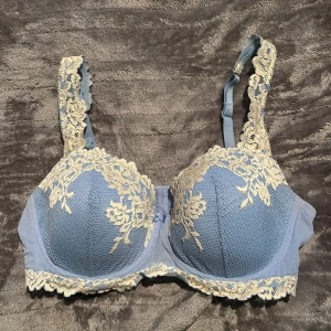 Blå spets-bh med broderade detaljer - Snygg blå bh från intimissimi med vackra broderade spetsdetaljer i vit längs kupor, axelband och nederkant. Kuporna är vadderade och har en liten rosett framtill. Justerbara axelband för perfekt passform. Perfekt för dig som gillar romantiska och feminina underkläder.