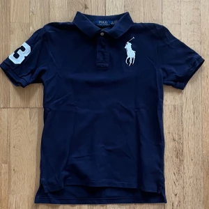 Marinblå pikétröja från Ralph Lauren - Marinblå pikétröja från Ralph Lauren med vit broderad logga på bröstet. OBS! Storlek 10-12 men sitter som Xs. Tveka inte på att höra av dig om du har några frågor eller funderingar!
