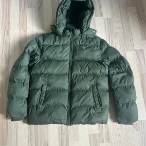 Grön pufferjacka från GANT med huva - Snygg mörkgrön pufferjacka från GANT med broderad logga på bröstet. Jackan har huva, dragkedja framtill och två fickor med knappstängning. Perfekt för kalla dagar och riktigt skön att bära. Klassisk quiltad look och stilren design.