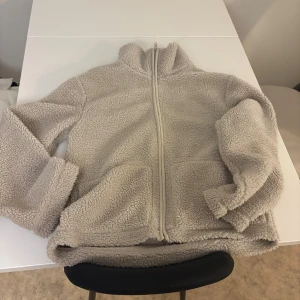 Beige teddyjacka från 157, storlek M - Mysig beige teddyjacka från 157 i storlek M. Jackan har hög krage, hel dragkedja framtill och två stora fickor. Perfekt för kalla dagar när du vill vara både varm och stilren. Fleece-liknande material som känns riktigt skönt mot huden. Lite smink upp vid kragen men går säkert att tvätta om man vill