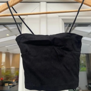 Svart ribbad linnetopp - Snygg svart ribbad linnetopp med smala axelband och croppad passform. Perfekt att styla med jeans eller kjol för en trendig look. Toppens material har en lätt struktur och känns mjuk mot huden.
