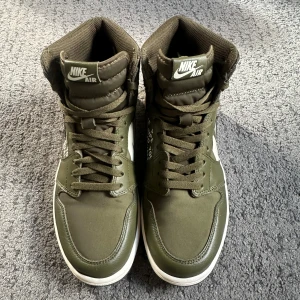 Nike Air Jordan 1 High Olive/White - Säljer ett par Nike Air Jordan 1 High i olivgrönt med vita detaljer och tryck på sidorna. Skorna har snörning, hög siluett och är tillverkade i skinn och syntet. Snygg kontrast mellan den gröna ovandelen och den vita sulan. Skorna som kommer inte till användning då de är lite för stora, skorna blivit använda några gånger och är i nästan nyskick. Storlek är 45 vilket motsvarar 44-45, ingen box följer med.  OBS INGEN FRAKT! Bara avhämtning i Göteborgsområdet. 