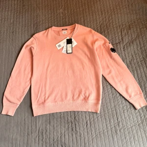 Rosa sweatshirt från C.P. Company - Snygg rosa sweatshirt från C.P. Company i bomull. Tröjan har rund hals, ribbade muddar och en ikonisk ficka med glaslins på ena ärmen. Perfekt för dig som gillar streetwear och vill sticka ut med en stilren look.
