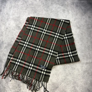 Rutig halsduk från Burberry i ull - Klassisk rutig halsduk från Burberry i mjuk ull. Färgerna är mörkgrön, svart, vit och röd med fransar i ändarna. Perfekt accessoar för att lyfta din outfit med en tidlös brittisk känsla.