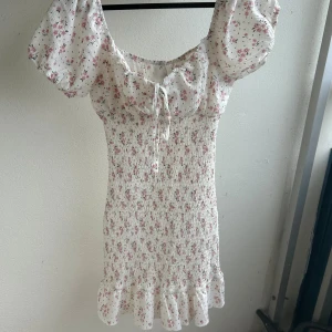Vit blommig klänning med puffärm - Supersöt vit klänning med rosa blommor , puffärmar som man kan sätta som off-shoulder oxå och volangkant nedtill. Linne tyg, väldigt stretchig i mitten av den som syns på bilden så den passar S-L men den är S. Jag är 169 cm och den går till strax över knäna på mig 