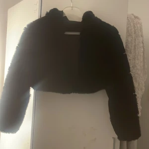 Svart croppad pälsjacka med huva - Säljer en svart, croppad pälsjacka med huva. Jackan har lång ärm och är gjord i mjukt fuskpälsmaterial som ger en lyxig och trendig vibe. Den håller dig varm under kyliga dagar. Passformen är normal och jackan är utan synliga knappar eller dragkedja. Den har 3smpa knappar som inte syns 