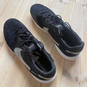 Nike Gato IC svart/vit sneakers 45 1/2 - Säljer ett par Nike Gato IC sneakers i svart med vit sula och silverfärgad swoosh. Skorna har ovandel i mesh och mocka, snörning och rund tå. Perfekta för inomhusfotboll eller streetstyle. Snygg och sportig design med tydliga Nike-detaljer.