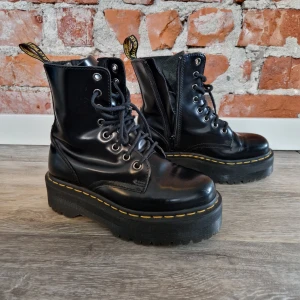 Dr. Martens Jadon svarta platåboots - Säljer ett par svarta Dr. Martens boots i storlek 36 med platåsula och klassiska gula sömmar. Skorna har snörning, dragkedja på sidan och är tillverkade i blankt skinn. Perfekta för dig som vill ha en edgy och cool stil.