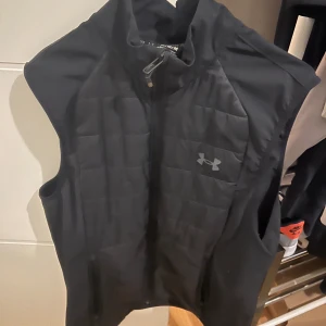 Svart dunväst från Under Armour - Sportig svart dunväst från Under Armour med quiltad framsida och dragkedja. Västen har hög krage och två fickor med dragkedja. Perfekt för lager-på-lager och har en clean look med diskret logga på bröstet.