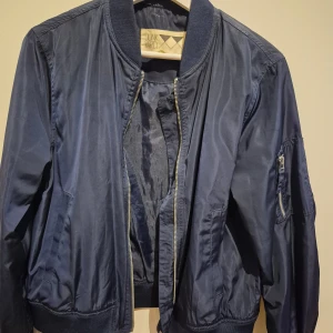 Mörkblå bomberjacka från Junkyard - Snygg mörkblå bomberjacka från Junkyard i klassisk modell med ribbade muddar, dragkedja framtill och ficka med dragkedja på ärmen. Jackan är tillverkad i polyester och har en stilren look som passar perfekt till streetwear.