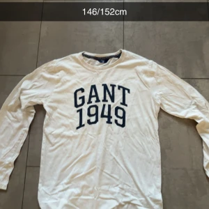 Vit långärmad tröja från Gant - Vit långärmad tröja från Gant med blå text 'GANT 1949' tryckt på bröstet. Klassisk rund halsringning och avslappnad passform. Perfekt för dig som gillar stilrena och sportiga plagg.