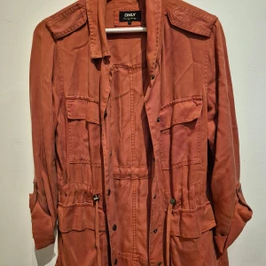Rostfärgad parkajacka från ONLY - Snygg rostfärgad parkajacka från ONLY med flera fickor, knappar och dragsko i midjan. Jackan har långa ärmar som kan rullas upp och fästas med knapp. Perfekt för dig som gillar en avslappnad och cool stil.